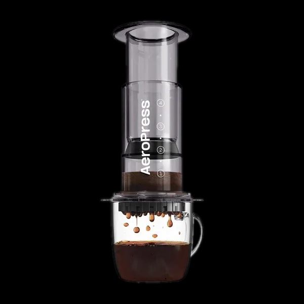 Zdjęcie czarnego zaparzacza AeroPress Clear Black, umieszczonego na szklance z zaparzaną kawą. Krople kawy spadają do kubka, ukazując proces parzenia metodą AeroPress, z przezroczystym czarnym korpusem urządzenia.