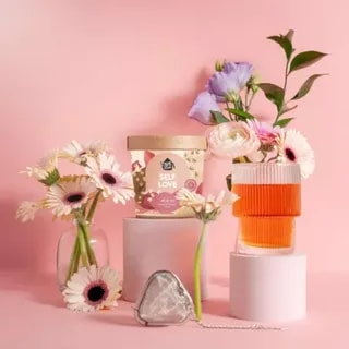 Na zdjęciu widoczna jest herbata "Self Love" w stylowym opakowaniu, otoczona kwiatami i akcesoriami do parzenia. Urokliwa aranżacja z różowymi tonami tworzy idealne tło dla produktów, które podkreślają miłość do siebie i przyjemność picia herbaty.