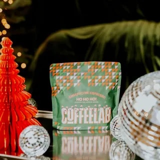 Pakowanie świątecznego espresso Ho Ho Ho! marki COFFEELAB, na którym widnieje kolorowa grafika z motywem świątecznym. W otoczeniu choinki i ozdób świetlnych, ten aromatyczny napój idealnie wpisuje się w bożonarodzeniowy klimat.