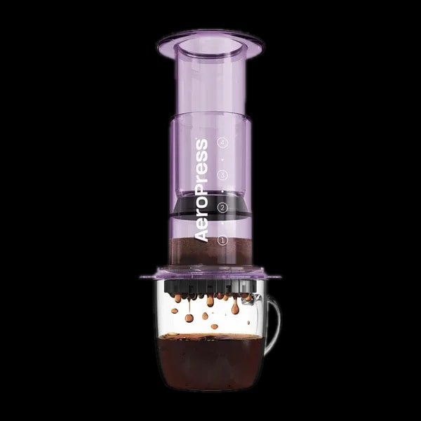 Zdjęcie fioletowego zaparzacza AeroPress Clear Purple, umieszczonego na szklance z zaparzaną kawą. Krople kawy spadają do kubka, ukazując proces parzenia metodą AeroPress, z przezroczystym fioletowym korpusem urządzenia.