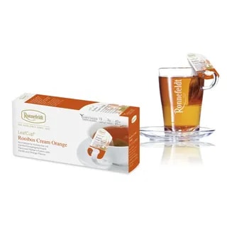 Opakowanie herbaty Ronnefeldt LeafCup Rooibos Cream Orange obok szklanki z przygotowaną herbatą. Herbata Rooibos Cream Orange to czarna herbata z mieszanką przypraw, oferowana w eleganckim pudełku z 15 torebkami herbaty, idealna do przygotowania aromatycznego napoju. Obok znajduje się przezroczysta szklanka z logo Ronnefeldt, ukazująca napój o bogatej, ciemnej barwie.