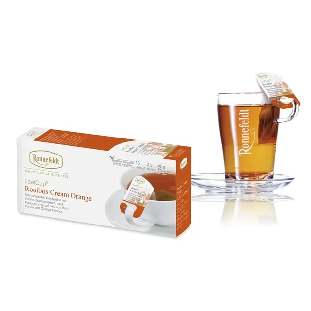 Opakowanie herbaty Ronnefeldt LeafCup Rooibos Cream Orange obok szklanki z przygotowaną herbatą. Herbata Rooibos Cream Orange to czarna herbata z mieszanką przypraw, oferowana w eleganckim pudełku z 15 torebkami herbaty, idealna do przygotowania aromatycznego napoju. Obok znajduje się przezroczysta szklanka z logo Ronnefeldt, ukazująca napój o bogatej, ciemnej barwie.