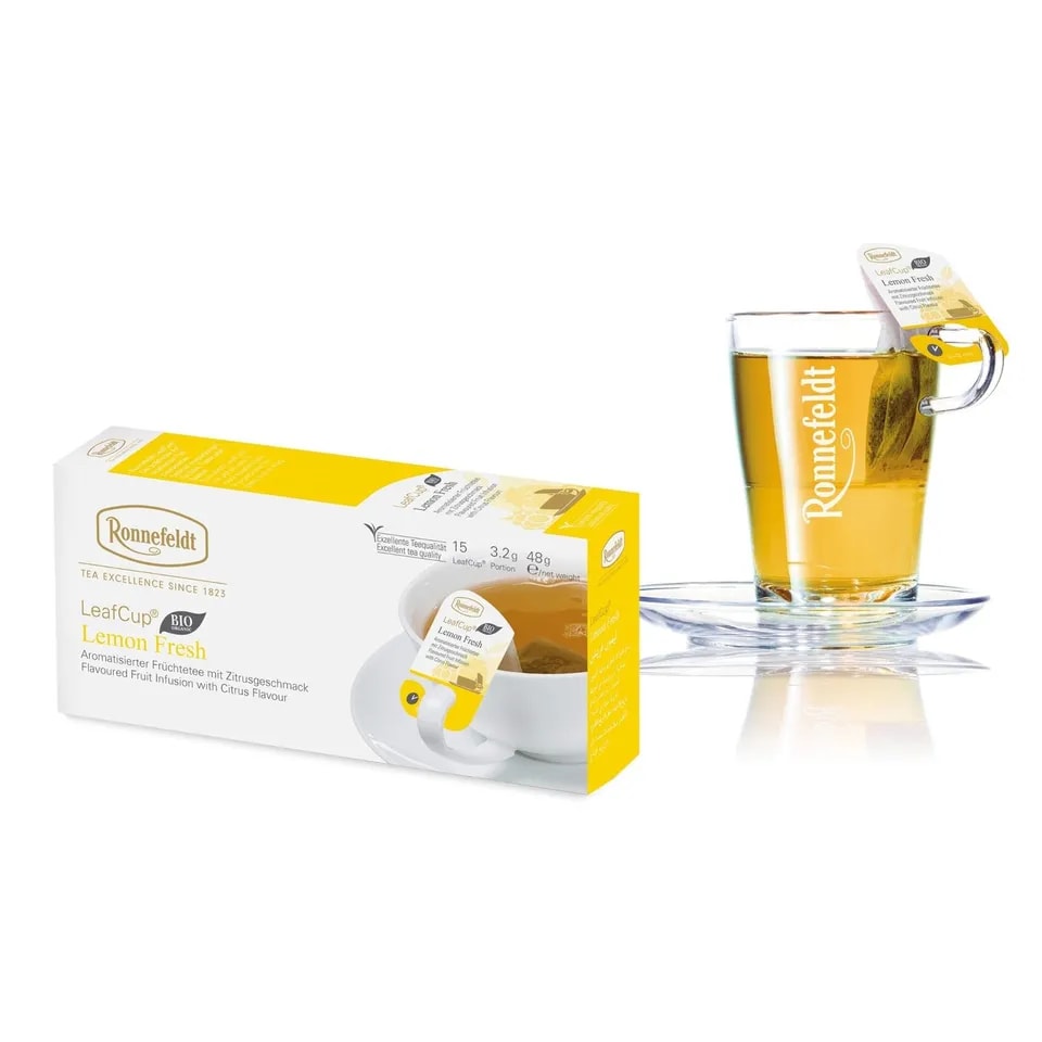 Opakowanie herbaty Ronnefeldt LeafCup Lemon Fresh obok szklanki z przygotowaną herbatą. Herbata Lemon Fresh to czarna herbata z mieszanką przypraw, oferowana w eleganckim pudełku z 15 torebkami herbaty, idealna do przygotowania aromatycznego napoju. Obok znajduje się przezroczysta szklanka z logo Ronnefeldt, ukazująca napój o bogatej, jasnej barwie.