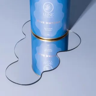 Opakowanie Blue Matcha od Lune Tea, z pięknym, niebieskim kolorem, znajduje się na lustrzanej powierzchni, co podkreśla jego unikalny design. Ten wysokiej jakości proszek z kwiatu Klitorii Ternateńskiej jest idealny do przygotowywania zdrowych napojów oraz deserów, dodając wyjątkowego smaku i koloru.