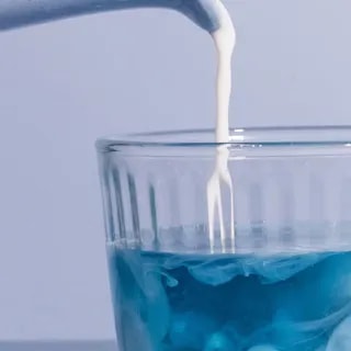 Obraz przedstawia mleko wlewające się do szklanki z niebieskim napojem, tworząc efektowne wzory. Doskonały sposób na przygotowanie herbaty lub innego napoju na bazie herbaty, idealny dla miłośników herbacianych eksperymentów.