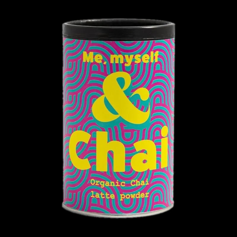 Kolorowe opakowanie Organic Chai Latte Powder marki Me, Myself & Chai. Cylindryczna puszka z intensywnym wzorem różowo-niebieskich fal oraz wyraźnym żółtym napisem 'Me, myself & Chai'. Zawiera organiczny proszek do przygotowania chai latte, idealny dla miłośników tej aromatycznej i korzennej herbaty.