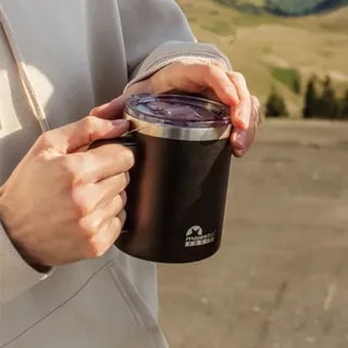 Osoba trzyma elegancki czarny kubek termiczny z metalową pokrywką, idealny do picia kawy lub herbaty. Kubek znajduje się w plenerze, co podkreśla jego funkcjonalność podczas outdoorowych przygód.
