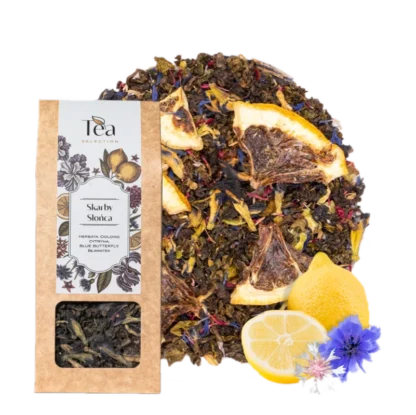 Opakowanie herbaty Skarby Słońca na białym tle, zawierającej mieszankę herbaty oolong, cytryny, blue butterfly oraz bławatera, obok talerz z suszonymi owocami i kwiatami, połowa cytryny, całe cytryny oraz niebieskie kwiaty chabra i białe kwiaty bławatera