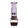 Zdjęcie fioletowego zaparzacza AeroPress Clear Purple, umieszczonego na szklance z zaparzaną kawą. Krople kawy spadają do kubka, ukazując proces parzenia metodą AeroPress, z przezroczystym fioletowym korpusem urządzenia.