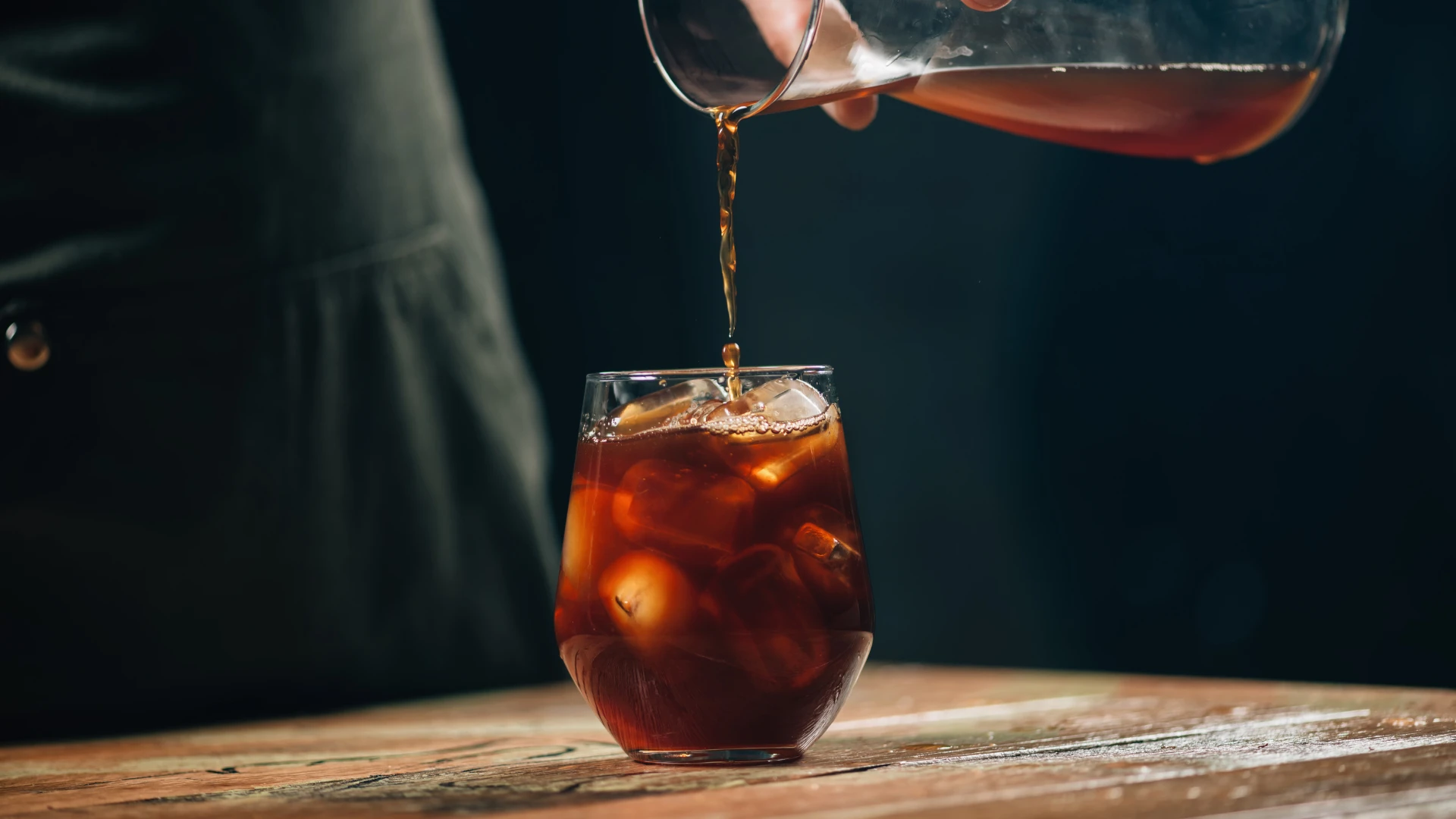 Kawa cold brew nalewana z szklanej karafki do szklanki wypełnionej kostkami lodu, stojącej na drewnianym stole.